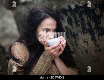 Natur, Schönheit, Jugend und gesundes Lifestyle-Konzept. Junge süße Mädchen in einem gestrickten Pullover vor einem Hintergrund einer alten Betonwand trinken Tee Fr. Stockfoto