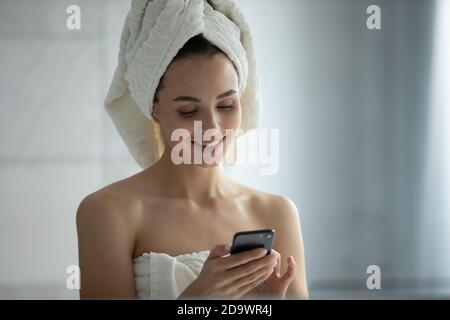 Lächelnde junge Frau, die nach der Dusche mit dem Handy im Badezimmer stand Stockfoto