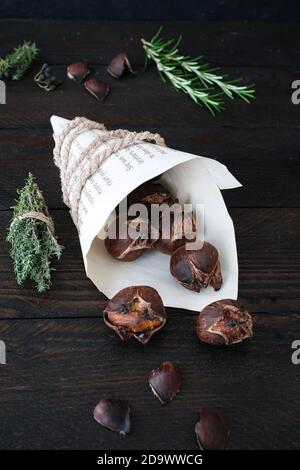 Frisch geröstete Kastanien, eingewickelt in einen Zeitungsbeutel, mit einer dicken Kordel gehalten, hübsch dekoriert auf einem dunklen Holztisch Stockfoto