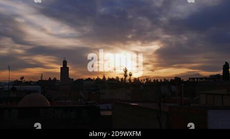 Dramatischer Morgenhimmel mit heller Sonne über dem historischen Zentrum (Medina) von Marrakesch, Marokko mit Silhouetten von Dächern, Palmen und einem Minarett. Stockfoto