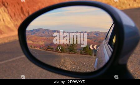 Wunderschön rot gefärbte Ausläufer des atemberaubenden Atlasgebirges in der Abendsonne mit gepflasterten Straßen und Straßenschildern im Seitenspiegel des Autos gesehen. Stockfoto