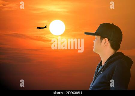 Asiatischer Mann trägt ein blaues Langarm-Shirt und trägt einen Hut Blick auf das Flugzeug am roten Himmel mit schönen Sonnenlicht. Stockfoto