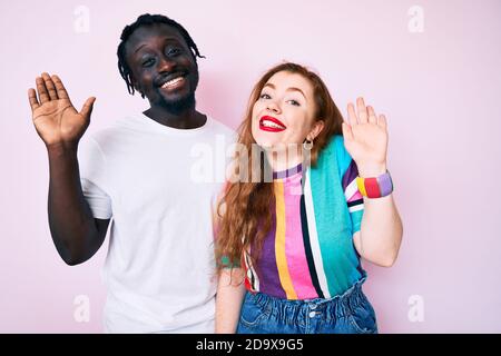 Interracial paar in legerer Kleidung waiving sagen Hallo glücklich und lächelnd, freundliche Willkommensgeste Stockfoto