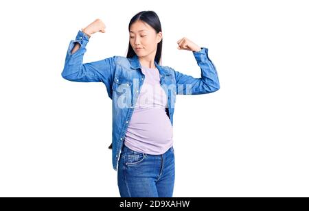 Junge schöne chinesische Frau schwanger erwartet Baby zeigt Arme Muskeln lächelnd stolz. Fitness-Konzept. Stockfoto