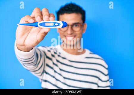 Junge gut aussehende hispanische Mann hält Thermometer positiv und glücklich Stehen und lächeln mit einem selbstbewussten Lächeln, das Zähne zeigt Stockfoto