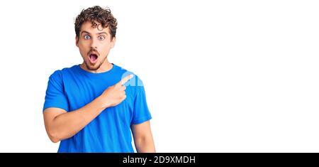 Junger hübscher Mann mit lockigem Haar trägt Freizeitkleidung überrascht zeigt mit Finger zur Seite, offenen Mund erstaunt Ausdruck. Stockfoto