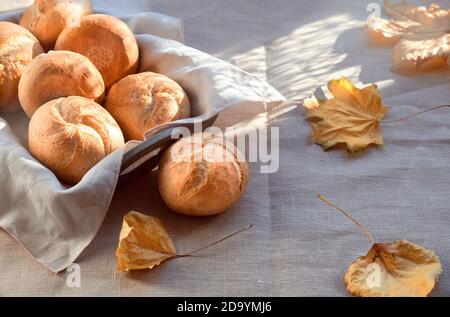 Kaiser oder Wien Brötchen im Brotkorb mit Handtuch. Tisch bedeckt mit beigefarbener Tischdecke. Orangefarbene Herbstblätter. Lichter mit langen Schatten. Stockfoto
