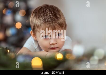 Ein schöner Junge von 4-5 Jahren sitzt oder steht in der Nähe des Weihnachtsbaums. Er träumt von einem Urlaub und Geschenke, macht einen Wunsch. Das Konzept des wi Stockfoto