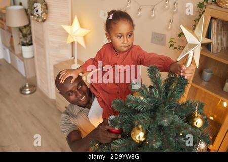 Hochwinkel-Porträt von afroamerikanischen Mädchen setzen Stern auf Weihnachten Baum mit Vater hilft ihr während der Weihnachtszeit zu genießen Zu Hause Stockfoto