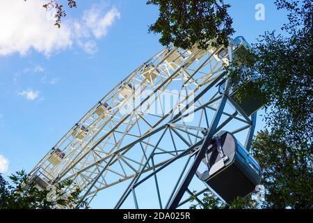 Riesenrad durch grünen Baum auf dem Hintergrund des blauen Himmels in sonnigen Sommertag, Ansicht von unten. Stockfoto