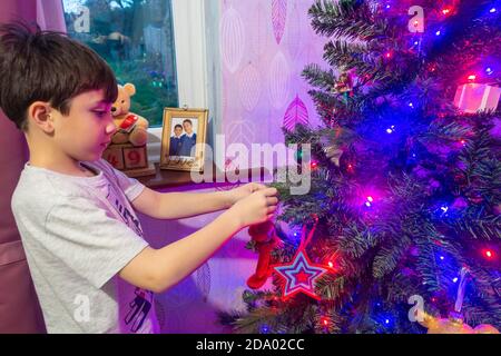 Ein Kind hängt Dekorationen auf einem Weihnachtsbaum, der y farbige Lichterketten beleuchtet ist. Stockfoto