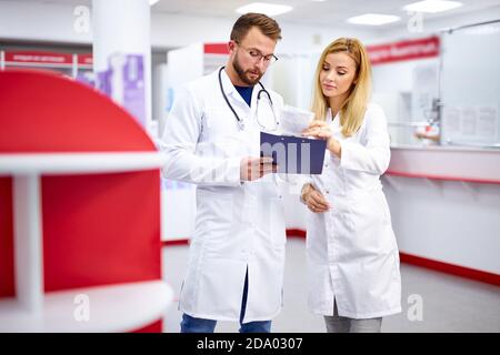 Zwei junge kaukasische Apotheker diskutieren Medikamente, mit einer digitalen Tablette und Dokumente. Mann und Frau in weißem medizinischen Kleid arbeiten in der Apotheke Stockfoto