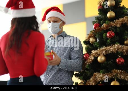 Junger Mann mit weihnachtsmann Hut und Schutzmaske auf Gesicht Geschenk an Frau in der Nähe weihnachtsbaum Stockfoto