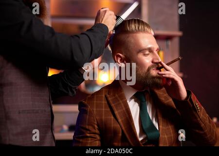 Stilvoller Mann genießen Frisur beim Friseur, er raucht während des Schneidens, Friseur schneidet die Haare auf dem Kopf des Kunden, während er Zigarette raucht, trägt formelle Kleidung Stockfoto