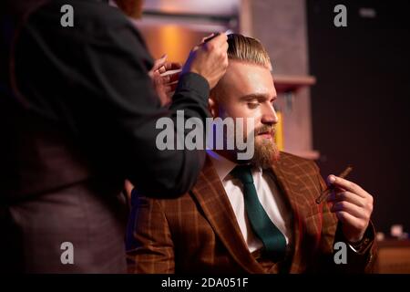 Stilvoller Mann genießen Frisur beim Friseur, er raucht während des Schneidens, Friseur schneidet die Haare auf dem Kopf des Kunden, während er Zigarette raucht, trägt formelle Kleidung Stockfoto