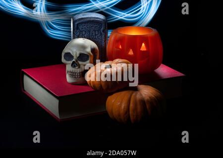 Halloween-Dekorationen und Miniatur-Kürbisse auf einem roten Buch isoliert In schwarzem Hintergrund und beleuchtet mit beweglichen blauen LED-Taschenlampe Stockfoto