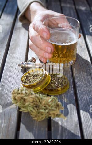 Nahaufnahme des Holztisches mit Marihuana-Mühle, Marihuana-Knospe, einer Rauchpfeife und einer männlichen Hand, die ein Glas Bier im Hintergrund hält. Stockfoto