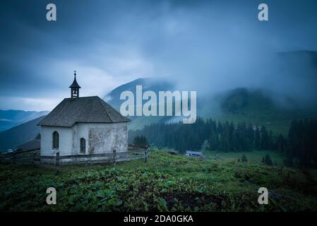 Weiße, mit Schindeldach bedeckte Kapelle auf den Ackernalmwiesen vor einem Bergpanorama mit bewölktem Himmel und Nebelwellen, Tirol, Österreich Stockfoto