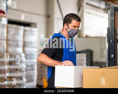 Seitenansicht Mitarbeiter der Logistikfirma in blauer Weste und Gesichtsmaske, die beim Arbeiten in der Nähe großer Kartonbehälter steht Geräumiges Lager Stockfoto