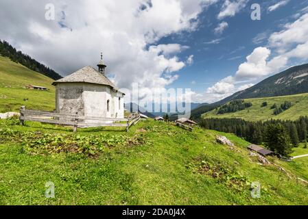 Weiße, mit Schindeldach bedeckte Kapelle auf der Ackernalm Almwiesen vor einem Bergpanorama in Sonnenschein, Tirol, Österreich Stockfoto