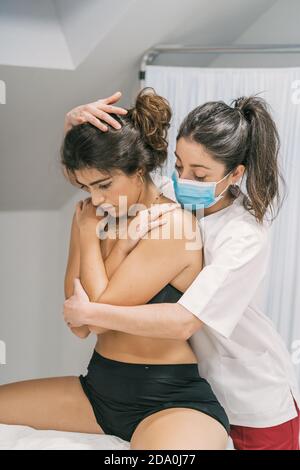 Seitenansicht der weiblichen Physiotherapeutin in medizinischer Maske und weiß Kittel berühren Kopf und Körper des Patienten und helfen während Therapiesitzung Stockfoto