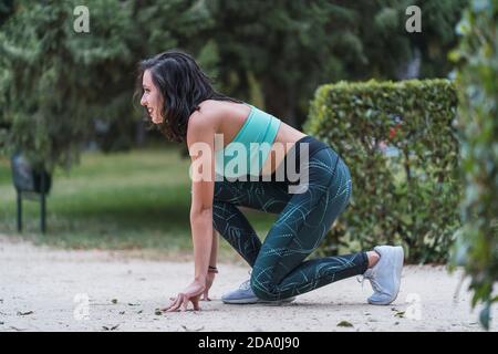 Seitenansicht einer lächelnden Läuferin, die im Crouch-Start steht Position und Vorbereitung für den Lauf während des Trainings im Sommer parken Stockfoto