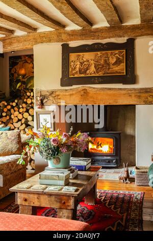 Interieur eines englischen Cottage mit beleuchteten Holzofen und Blumenschmuck in einer komfortablen Lounge mit freiliegenden Deckenbalken. Stockfoto