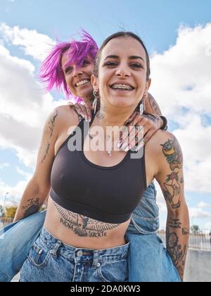 Niedriger Winkel der entzückten Frau mit rosa Haaren Huckepack fröhliche Freundin mit Tattoos, während Spaß zusammen am Wochenende Stockfoto