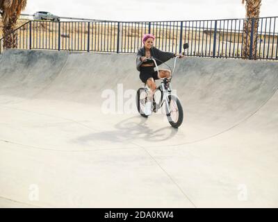 Tätowierte Frau in stilvollem Outfit sitzt auf BMX Fahrrad in Skatepark und wegschauen Stockfoto