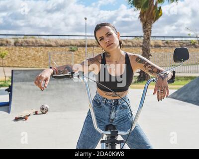Bestimmt tätowierte Frau in stilvollem Outfit auf BMX Fahrrad sitzen Im Skatepark und beim Blick auf die Kamera Stockfoto