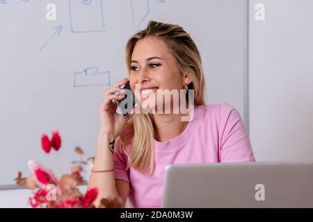 Entzückte Freiberuflerin diskutiert Projekt auf Smartphone während sitzen Tisch mit Laptop und Remote-Arbeit zu Hause Stockfoto