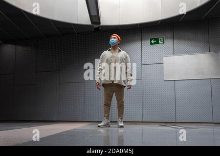 Ganzkörper konzentriert Stylistin asiatische Mann tragen lässige Outfit und Gesichtsmaske, die auf dem Flur eines Betongebäudes steht und wegschaut Stockfoto