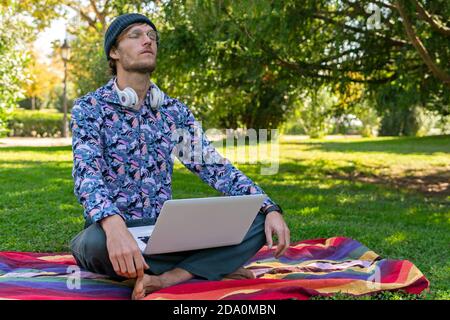 Calm männlichen Unternehmer sitzen im Park mit Laptop und tun Yoga in Lotus Pose mit geschlossenen Augen während der Pause Bei Remote-Arbeit Stockfoto