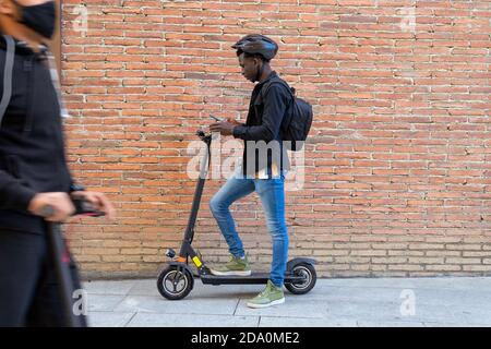 Seitenansicht des nachdenklichen afroamerikanischen Mannes, der auf elektrisch steht Scooter auf Bürgersteig und Surfen Smartphone während der Fahrt zur Arbeit Stockfoto