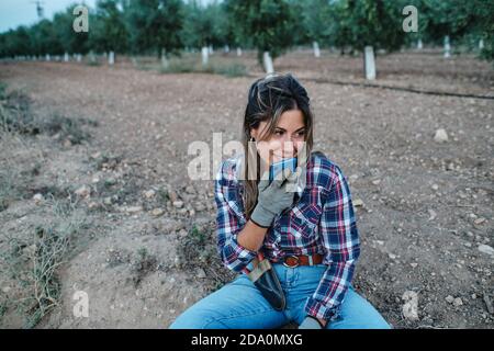 Bäuerin sitzt auf dem Boden und Aufzeichnung von Audio-Nachricht über Mobiltelefon während Sie sich während der Arbeit entspannen und wegschauen Stockfoto