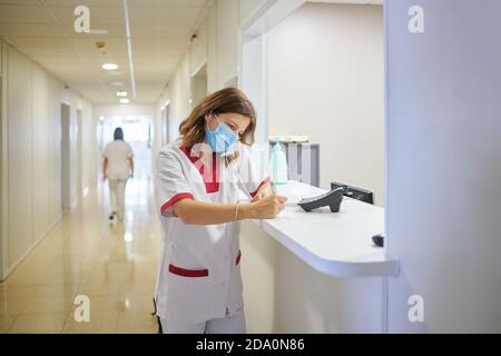 Konzentrierte weibliche Krankenschwester trägt weißen medizinischen Anzug und Gesichtsmaske Notizen in der klinischen Dokumentation machen, während man sich in der Nähe von modern befindet Krankenhausempfang Stockfoto