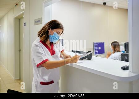 Konzentrierte weibliche Krankenschwester trägt weißen medizinischen Anzug und Gesichtsmaske Notizen in der klinischen Dokumentation machen, während man sich in der Nähe von modern befindet Krankenhausempfang Stockfoto