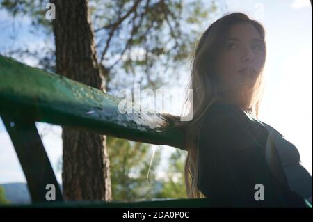 Besinnliche junge Frau in legerer Kleidung auf Holzbank sitzend Und genießen Sie die Sonne, während Sie sich im grünen Wald ausruhen Stockfoto