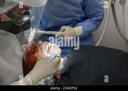 Professioneller Stomatologe mit Assistent, der Schutzmasken und Gesichtsschilde trägt Sitzen in der Nähe von Patienten mit Expander im Mund in der modernen Klinik Stockfoto