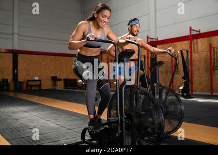 Fröhliche Sportlerin und Sportler radeln auf stationären Airbikes während des aktiven Trainings im modernen Fitnessstudio Stockfoto