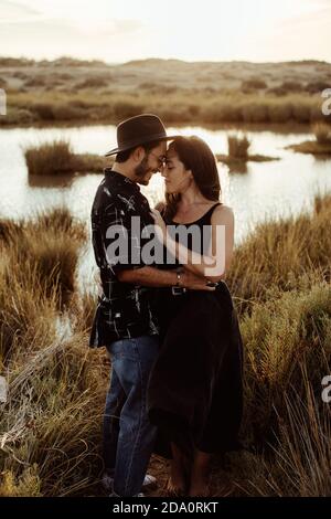 Ganzkörper Seitenansicht der jungen liebevollen Paar in casual Kleidung Kuscheln mit geschlossenen Augen, während auf Küste von stehen Reservoir umgeben von trockenem Gras i Stockfoto