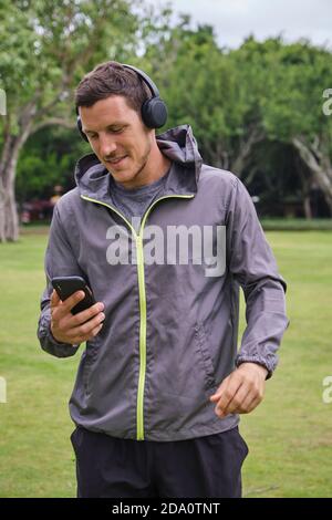 Lächelnder Läufer mit aktivem und kabellosem Kopfhörer, der Lieder auswählt Von der Playlist auf dem Smartphone während der Vorbereitung auf das Training im Park Stockfoto