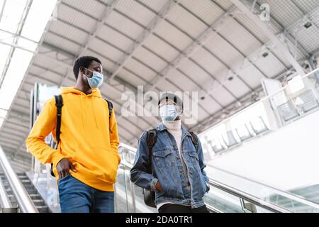 Niedriger Winkel der afroamerikanischen männlichen Freunde tragen medizinische Masken Auf der Rolltreppe auf dem modernen Bahnhof stehen und sich anschauen Einander Stockfoto