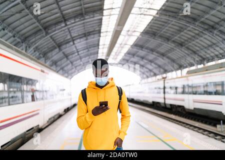 Ethnischer männlicher Reisender mit Rucksack und in Maske stehend Bahnsteig mit Zug und Handy während der Wartezeit auf die Abfahrt Während der COVID 19 epidemii Stockfoto