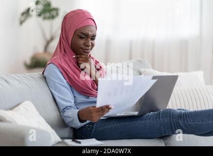 Nachdenkliche schwarze muslimische Frau, die Papiere überprüft und am Laptop arbeitet Zu Hause Stockfoto