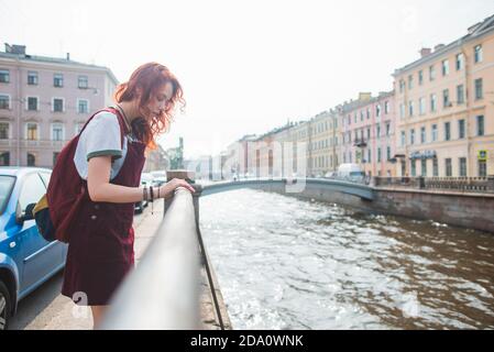 Seitenansicht der jungen rothaarigen weiblichen Reisenden in ungezwungener Atmosphäre Outfit mit Rucksack in der Nähe von Kanal ruhen, während die Erkundung der alten Architektur Von Sankt Petersburg Stockfoto