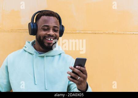Positive Erwachsene bärtigen afroamerikanischen Mann in Kapuzenpullover hören Musik mit kabellosen Kopfhörern und Smartphone-Browsing, während Sie dagegen stehen Brüllt Stockfoto