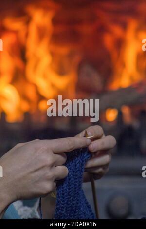 Vertikale Ansicht der Hände einer Frau, die im Hintergrund mit einem brennenden Kamin strickt. Stockfoto