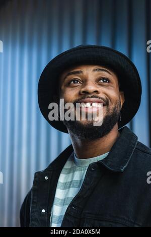 Happy African American bärtig Kerl tragen trendige Casual Outfit und hut lächelt hell und schaut nach oben, während sie gegen Shuttered steht Wand Stockfoto