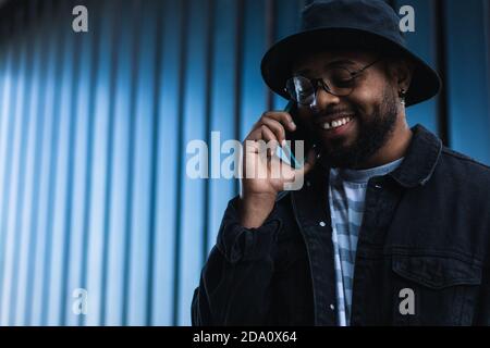 Fröhlicher afroamerikanischer Hipster-Rüde mit Hut und Brille Sprechen auf dem Handy, während Sie in der Nähe von Metallwand auf stehen Straße Stockfoto